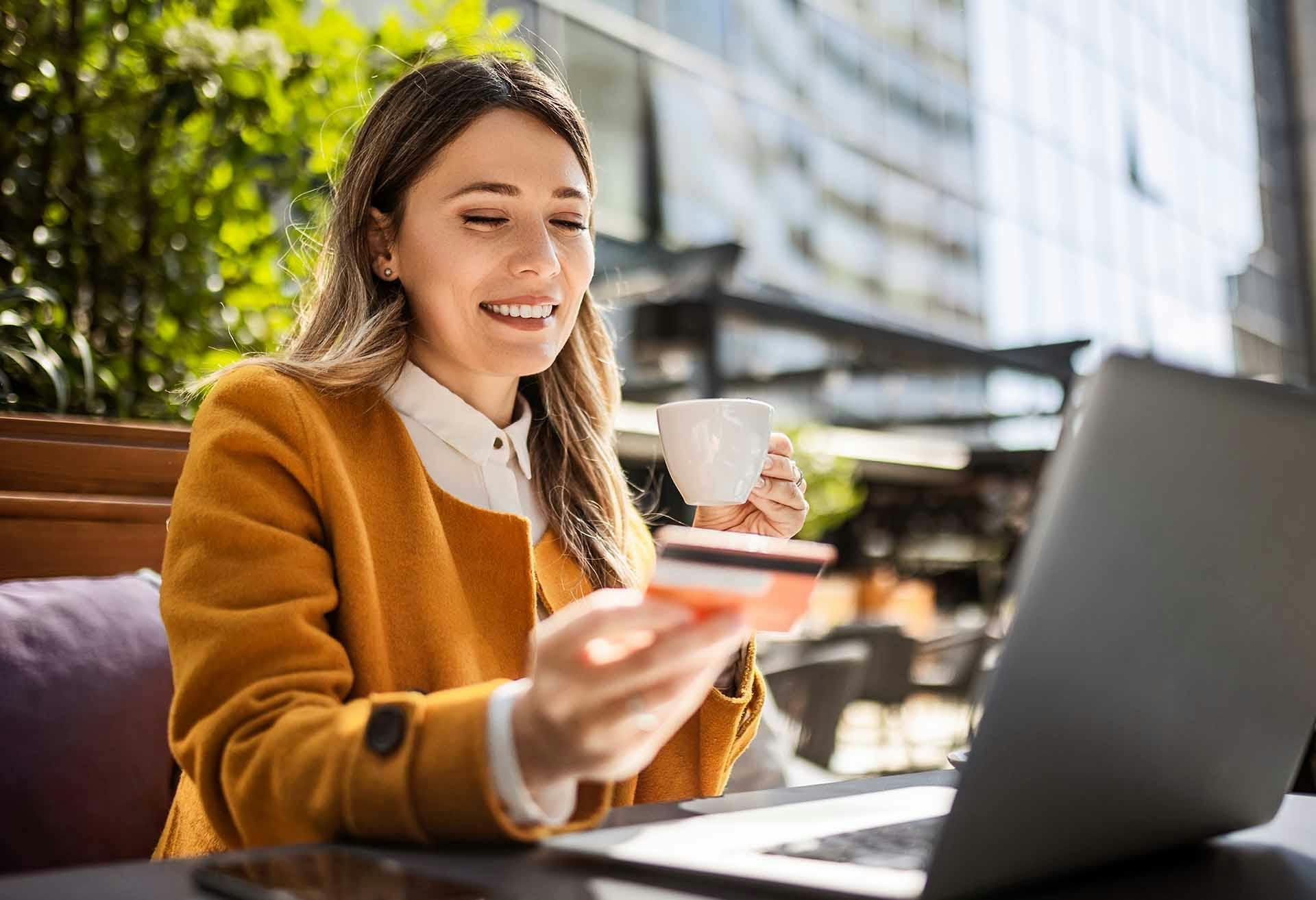 Woman with a cup in one hand, credit card in the other, in front of a laptop