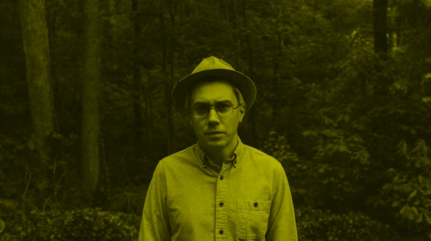 A person with light skin and short grey hair wearing a brimmed hat stands in shady a forest glade. They have a slightly concerned expression.