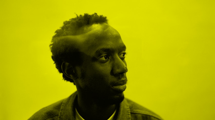 A person with short black hair and dark brown skin gazes to the right.