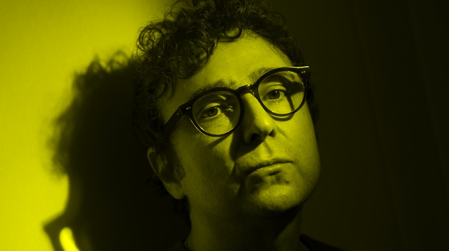A person with light skin and short, curly dark brown hair and thick-rimmed glasses leans against a wall. Their lips are pursed and they look serious.