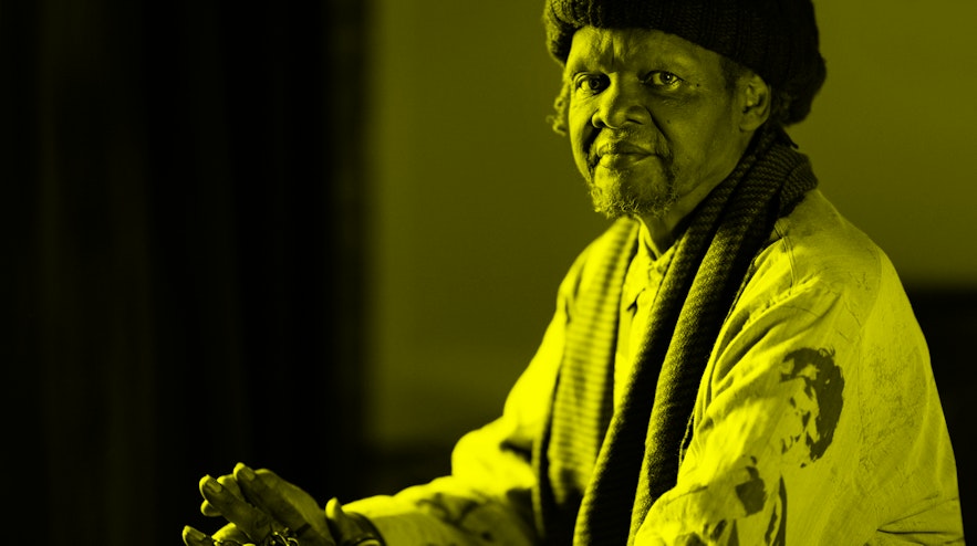 A person with medium brown skin and a light grey goatee sits with their hands pressed together. They have an even gaze.