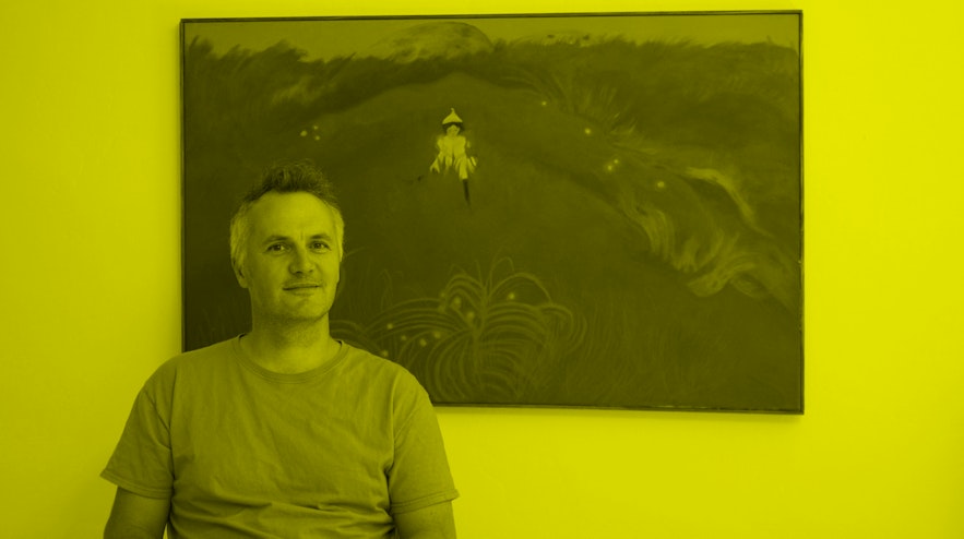 A person with short brown and white hair and light skin sits in front of a green painting. The are smiling.