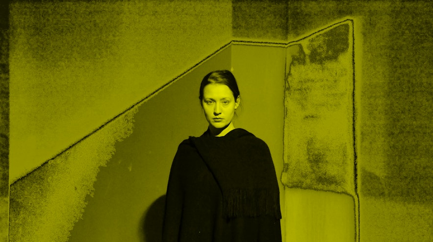 A person with light skin and long brown hair stands in a black gown. They are gazing at you with a neutral expression.