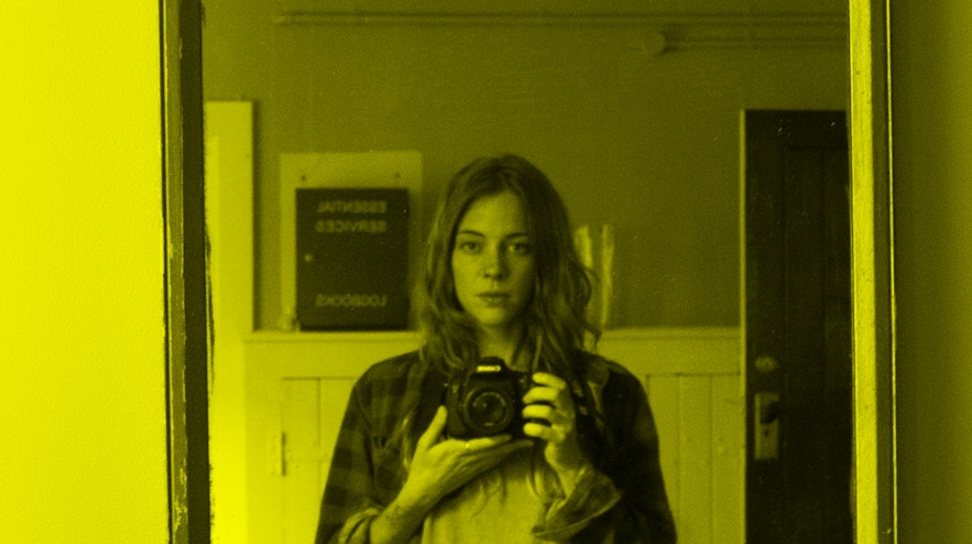A person with long red hair and light skin and a sullen expression taking a selfie in the mirror with a digital single lens reflex camera.