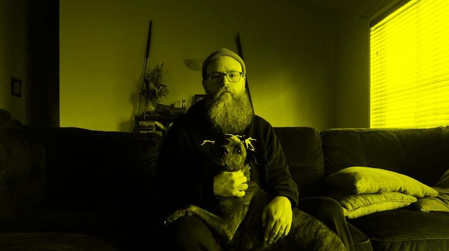 A person with a long red and grey beard and light skin with glasses sits on a sofa with a large brown dog in their lap. Both the person and the dog have serious expressions.
