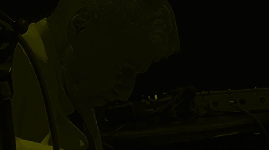A person with short white hair and light skin sits playing an electric guitar. Their head is bent forward in a look of concentration.