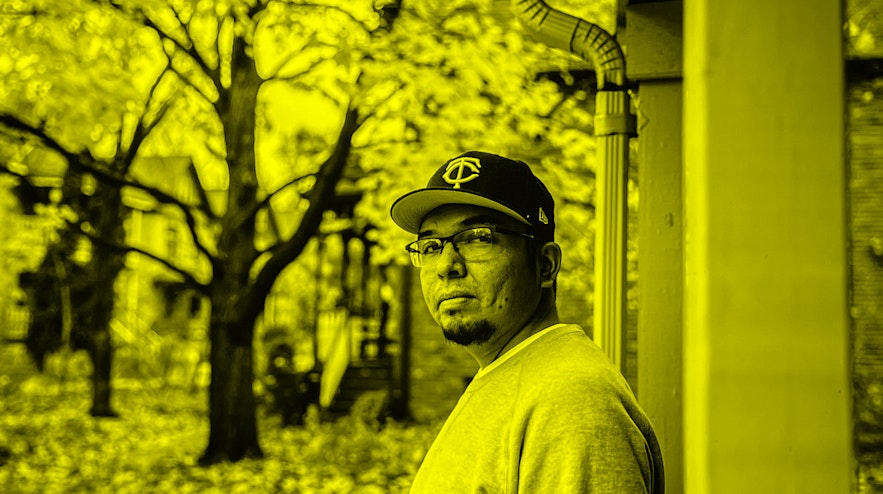 A person with light brown skin, a short goatee and glasses looks over their left shoulder with a thoughtful expression. Behind them are several autumn trees and a down pipe.