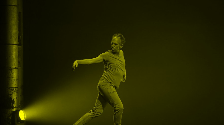 Person stepping across a stage with one leg behind and stretched, swinging both arms outward.