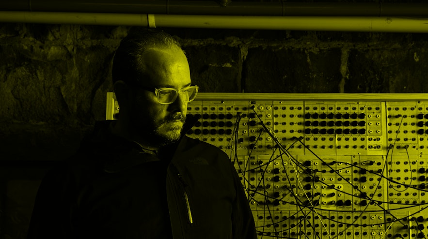 Person with dark hair and glasses, wearing all black, standing in front of a board covered in plugs and wires.