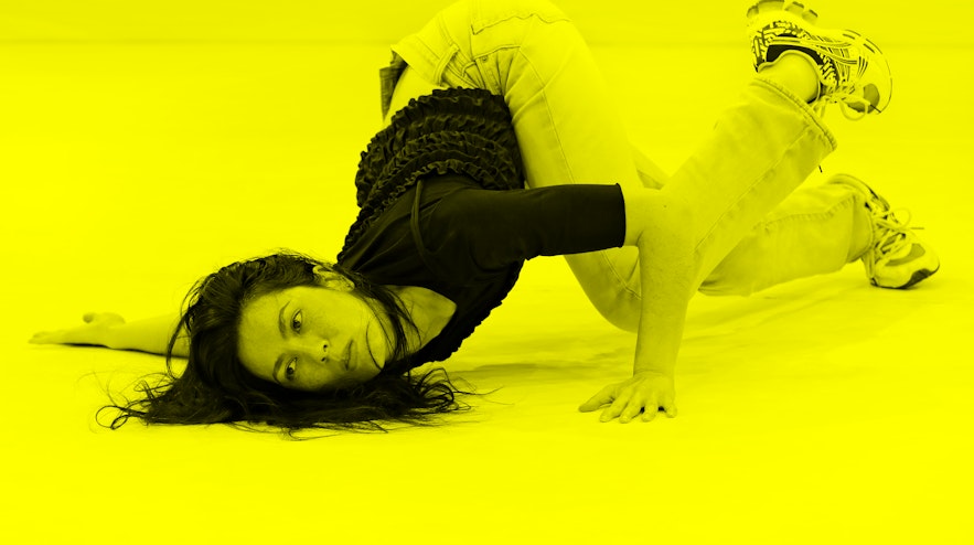 A person with long black hair and light skin contorts their body against the ground.