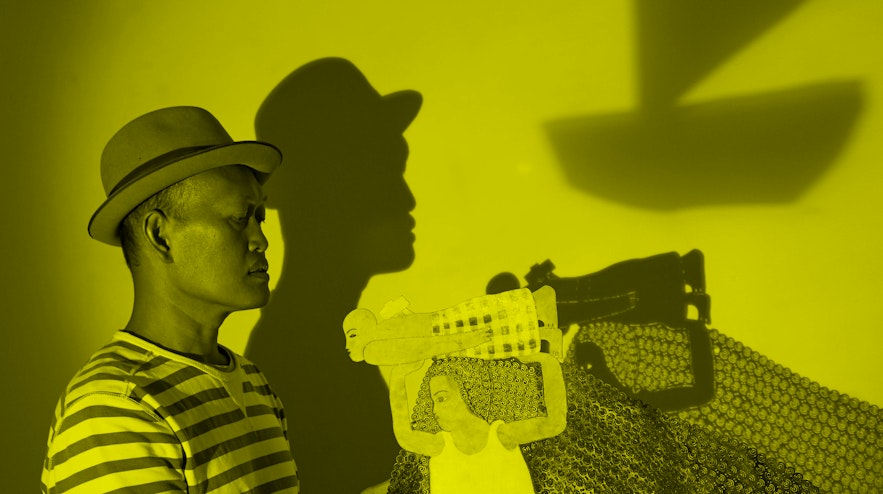 A person with medium brown skin and short black hair holds a paper cutout and net. The shadows are cast onto a wall behind them.
