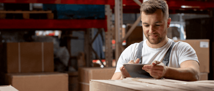 A man wearing overalls is standing in a warehouse, smiling while using a tablet. Large cardboard boxes are stacked around him, and shelves with more boxes are visible in the background.