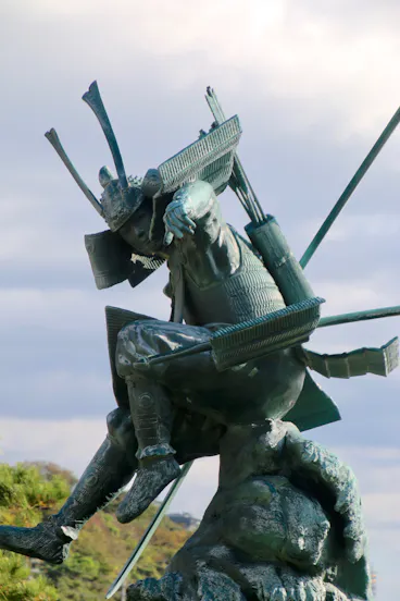 A bronze statue of a samurai warrior in traditional armor riding a horse, both in dynamic motion, with bows and arrows on the samurai’s back, set against a cloudy sky.