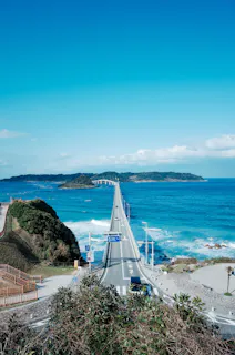 A long bridge stretches over bright blue ocean water, connecting the mainland to a distant green island under a clear, sunny sky. Signs, railings, and a few people are visible near the start of the bridge.