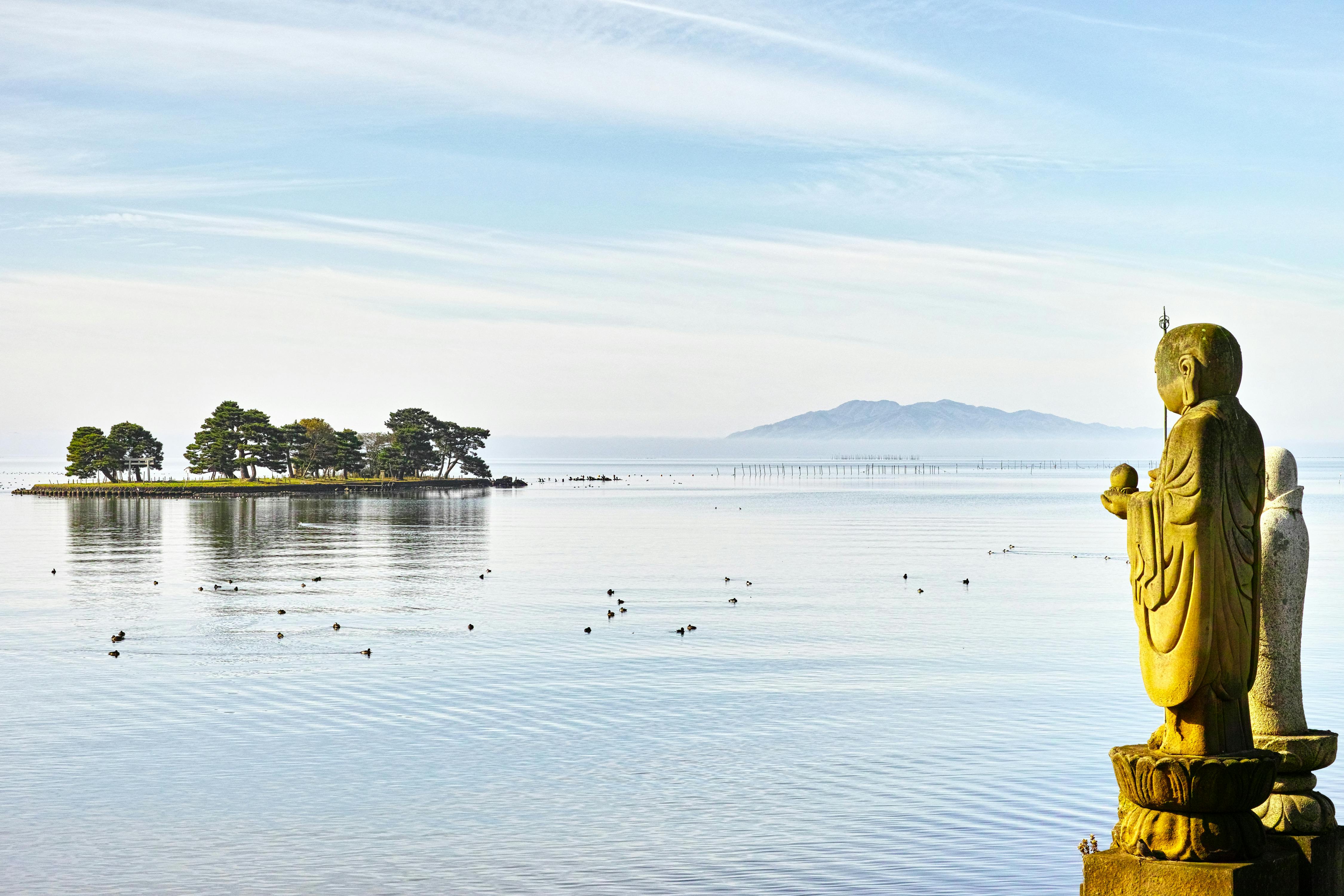A stone statue overlooks a calm lake dotted with birds, a small tree-covered island, and distant mountains beneath a blue, lightly clouded sky.