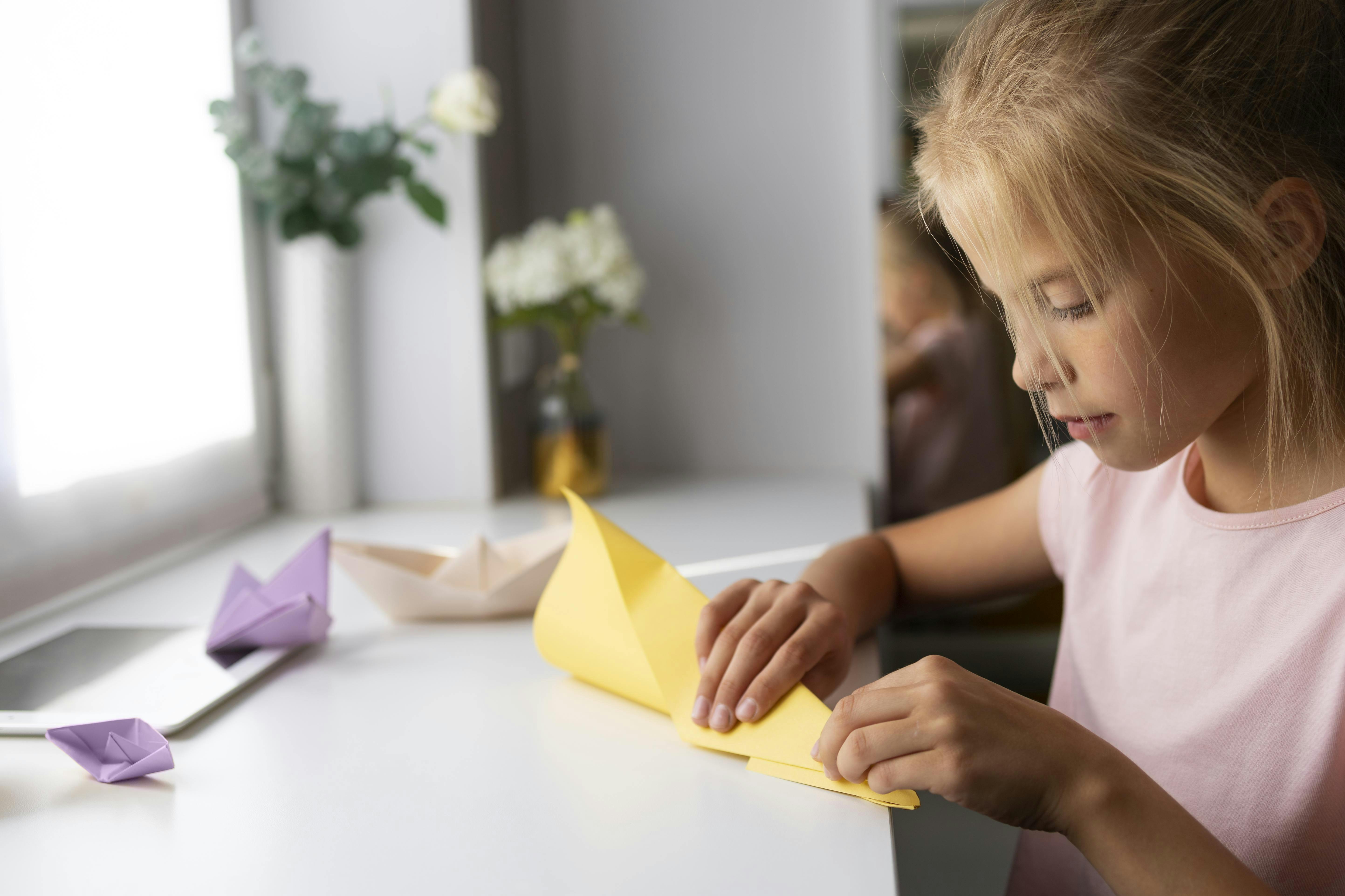 A young girl sits at a white desk, carefully folding a sheet of yellow paper. Origami boats in purple and beige rest nearby, and a vase with white flowers is in the background. Natural light comes in through a window.