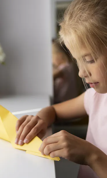 Girl doing origami A young girl sits at a white desk, carefully folding a sheet of yellow paper. Origami boats in purple and beige rest nearby, and a vase with white flowers is in the background. Natural light comes in through a window.