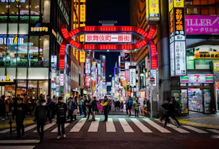 A busy street in Tokyo’s Kabukicho district at night, with a large red illuminated gate sign, crowds of people crossing, and colorful neon signs on tall buildings lining both sides.