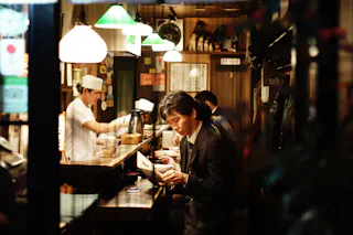 A man in a suit reads a menu at a cozy, warmly lit Japanese restaurant counter, while a chef in white prepares food in the background. The setting feels intimate and bustling.