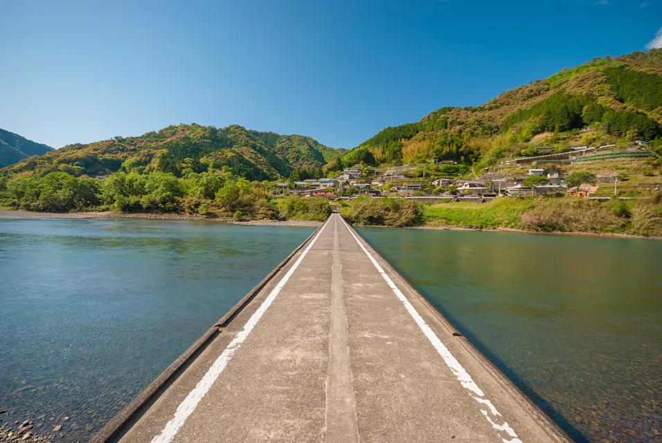 A narrow concrete bridge stretches across a wide, calm river leading towards a quaint village. The village is nestled among green, forested hills under a clear blue sky. The bridge has white lines painted on it and appears to be a pedestrian pathway.