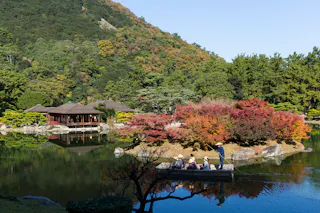 A serene Japanese garden with a pond, autumn-colored trees, a wooden building, and a small group of people on a boat near the shore, set against a lush, green mountain under a clear blue sky.