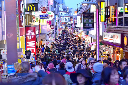 A busy street in Japan crowded with people, lined with brightly lit signs for shops and restaurants, including McDonald's and Wolfgang Puck, during daylight.