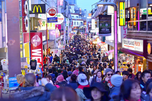 A busy street in Japan crowded with people, lined with brightly lit signs for shops and restaurants, including McDonald's and Wolfgang Puck, during daylight.