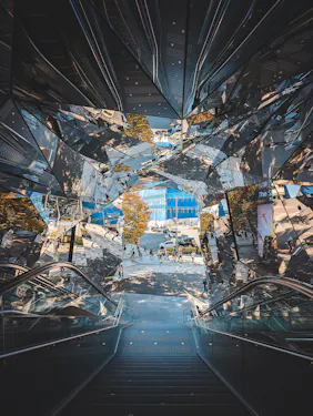 Glass-paneled escalators reflect trees, sky, and people outside a modern urban building, creating a kaleidoscopic effect as viewed from the top of the escalator looking down toward the street.