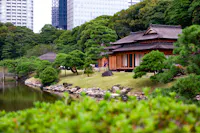 A traditional Japanese wooden building sits by a pond, surrounded by manicured trees and green shrubs, with modern skyscrapers visible in the background.