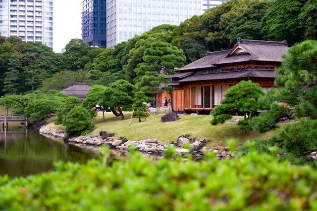 A traditional Japanese wooden building sits by a pond, surrounded by manicured trees and green shrubs, with modern skyscrapers visible in the background.