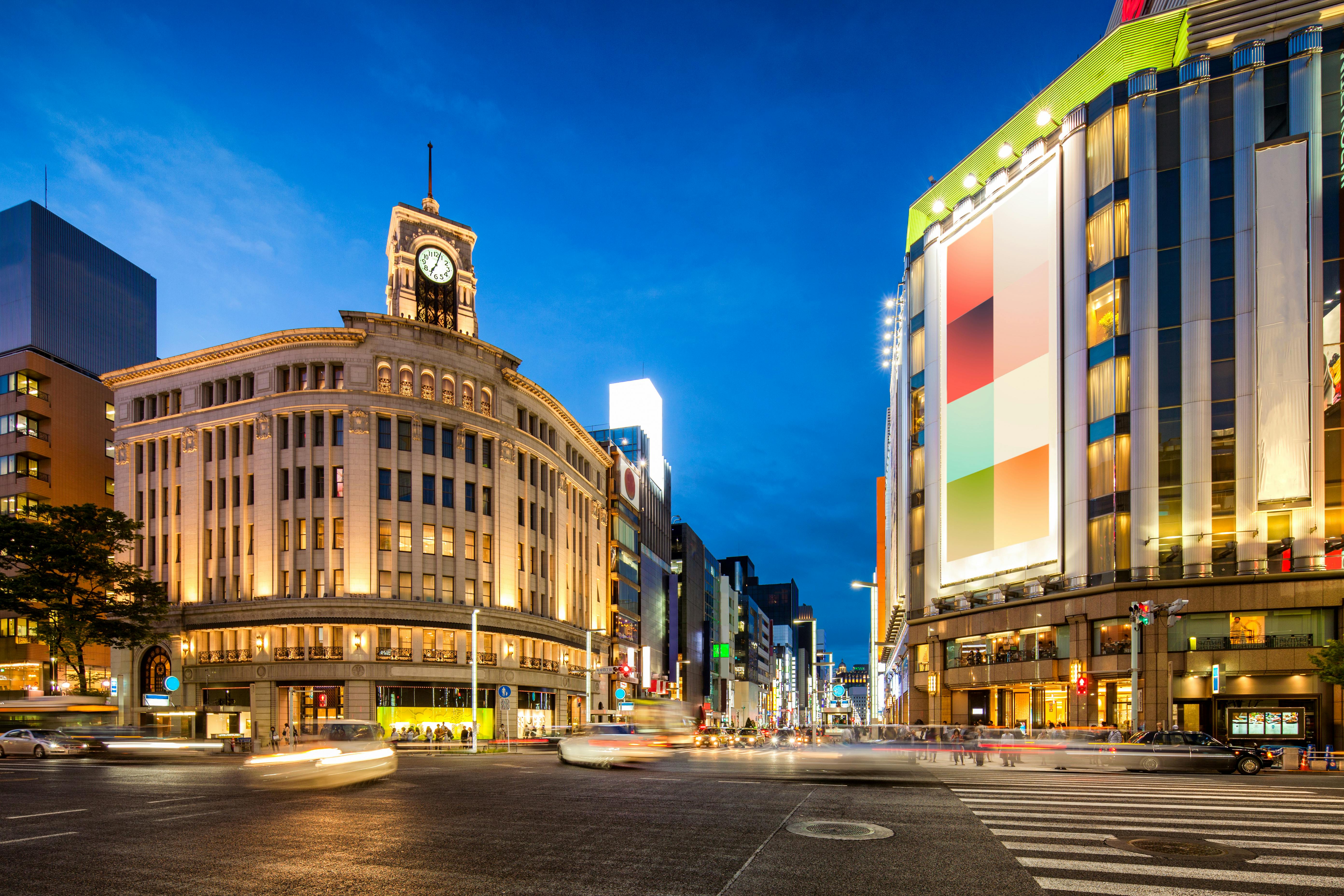 A brightly lit city intersection at dusk in Tokyo’s Ginza district, featuring the historic Wako clock tower on the left and modern buildings with illuminated signs and colorful billboards on the right.