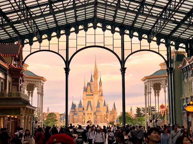 A large crowd gathers under an ornate glass and steel archway, with a fairytale castle in the background at sunset. The scene is lively, with people walking and taking photos in a theme park setting.