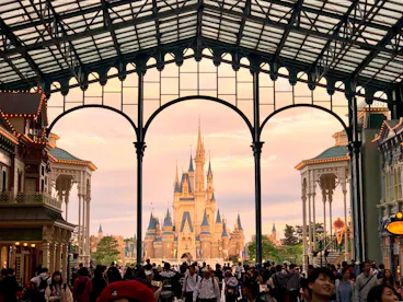 A large crowd gathers under an ornate glass and steel archway, with a fairytale castle in the background at sunset. The scene is lively, with people walking and taking photos in a theme park setting.