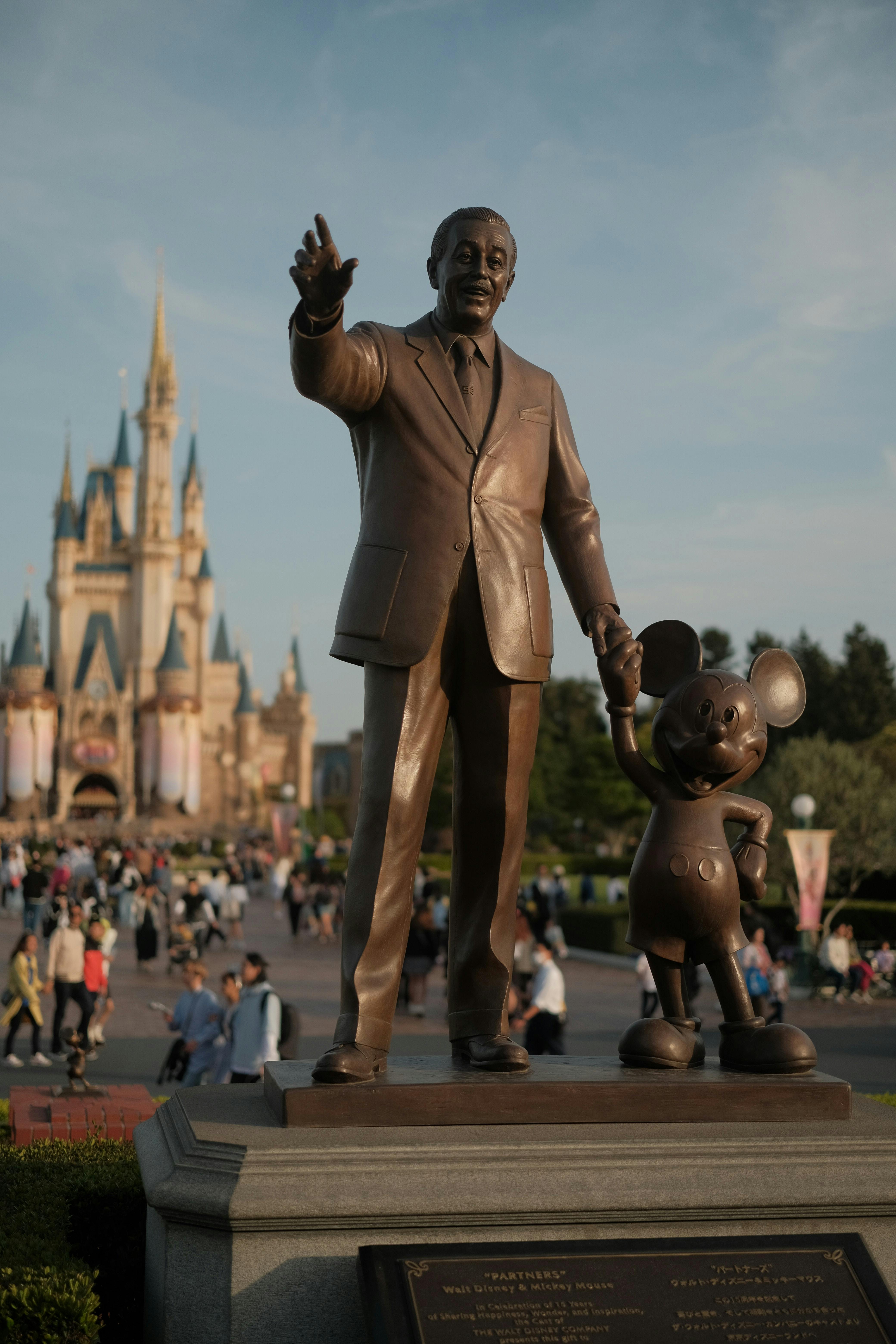 A bronze statue of a man holding hands with Mickey Mouse stands in front of a fairytale castle at a busy theme park, with visitors walking around and greenery in the background.