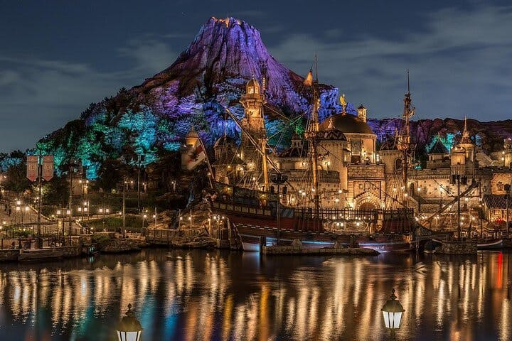 A brightly lit waterfront theme park at night, featuring a large, illuminated mountain in the background, ornate buildings, a sailing ship, and colorful lights reflected in the calm water.
