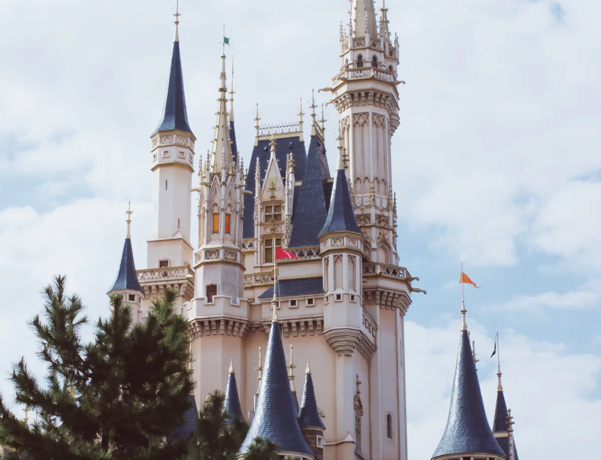 Disneyland A fairytale-style castle with tall spires and blue rooftops rises above green trees under a partly cloudy sky.