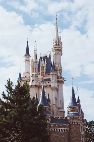 A fairytale-style castle with tall spires and blue rooftops rises above green trees under a partly cloudy sky.