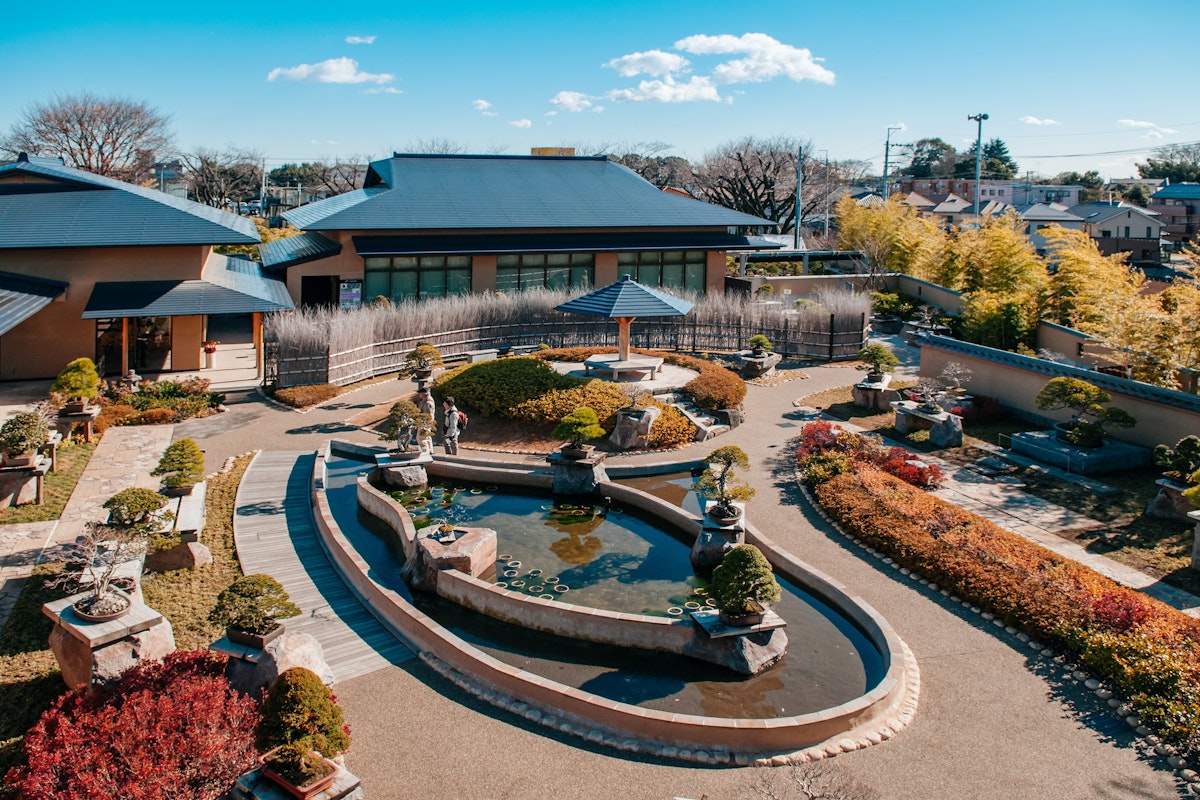 Omiya Bonsai Art Museum A serene Japanese garden with a central pond surrounded by neatly trimmed bushes and bonsai trees. The garden features winding pathways, miniature bridges, and stone lanterns. Traditional buildings with tiled roofs frame the scene under a bright blue sky.