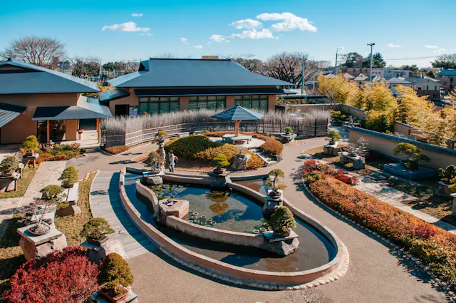 A serene Japanese garden with a central pond surrounded by neatly trimmed bushes and bonsai trees. The garden features winding pathways, miniature bridges, and stone lanterns. Traditional buildings with tiled roofs frame the scene under a bright blue sky.