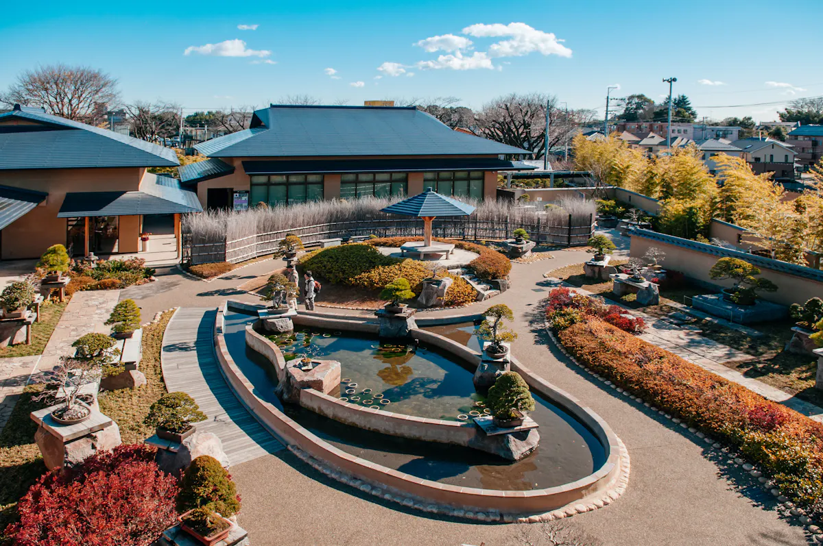 A serene Japanese garden with a central pond surrounded by neatly trimmed bushes and bonsai trees. The garden features winding pathways, miniature bridges, and stone lanterns. Traditional buildings with tiled roofs frame the scene under a bright blue sky.