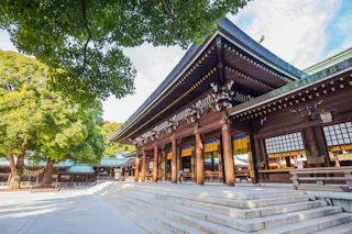 A traditional Japanese Shinto shrine with wooden architecture, tall pillars, and a sloping roof. Large green trees surround the shrine, and stone steps lead up to the entrance on a bright, sunny day.