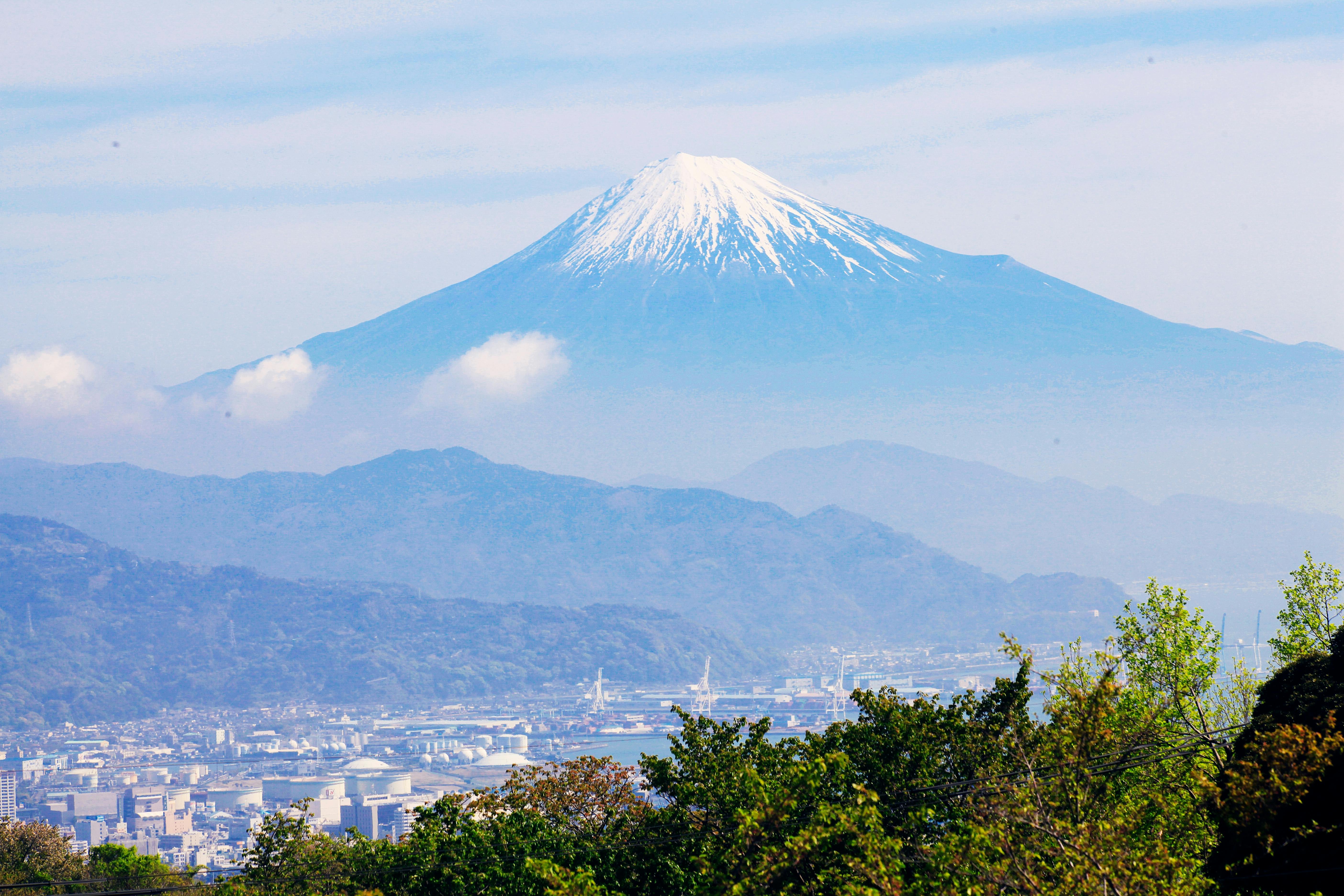 Snow-capped Mount Fuji rises above a cityscape, framed by green trees in the foreground and rolling hills under a blue sky with light clouds.