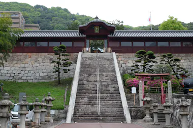 Wide stone steps lead up to a traditional Japanese-style wooden building with a tiled roof, surrounded by manicured trees, stone lanterns, and colorful shrubs, set against a backdrop of green hills.