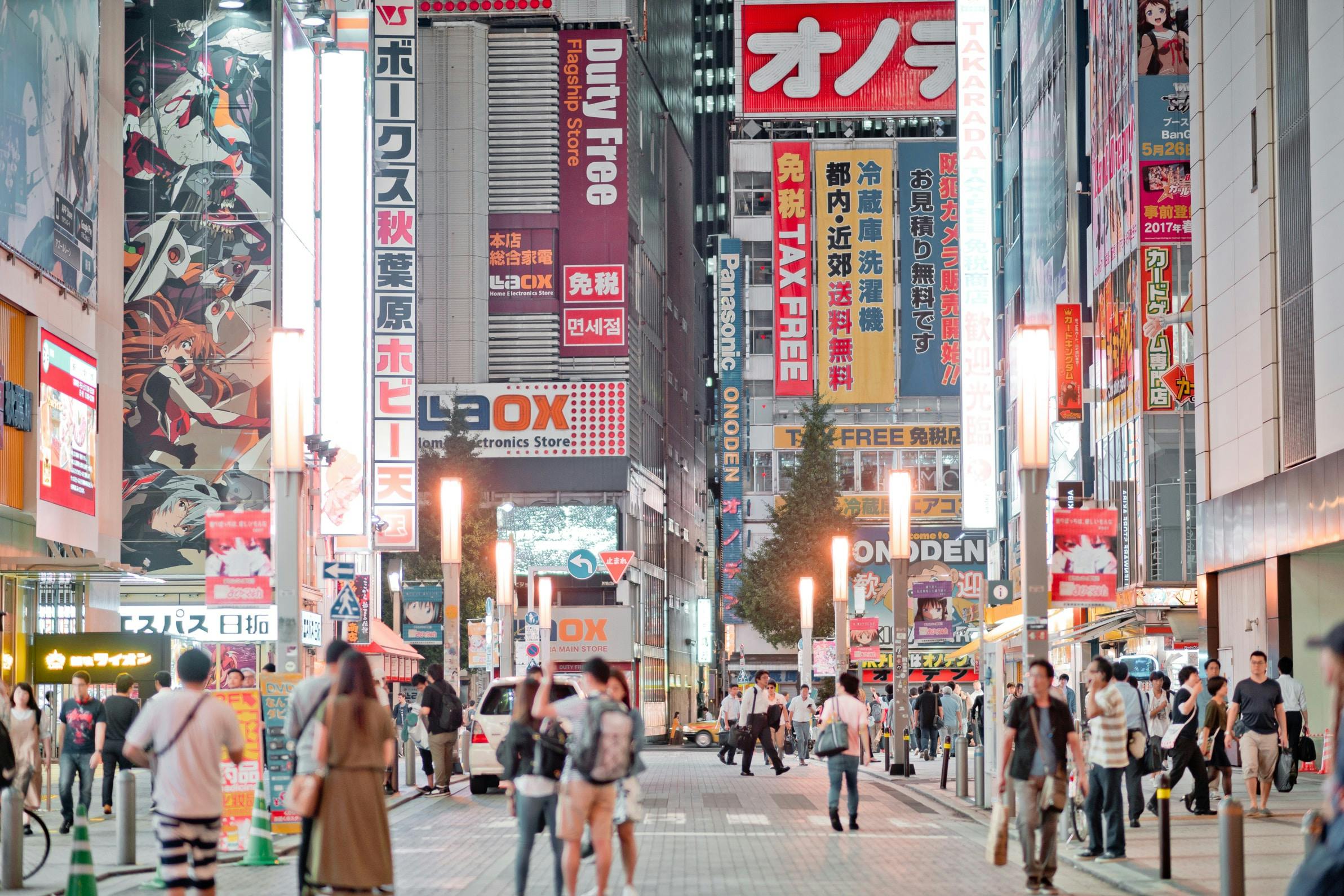 A busy street in a Japanese city at night, filled with people walking and colorful neon signs and advertisements on tall buildings, creating a vibrant urban atmosphere.