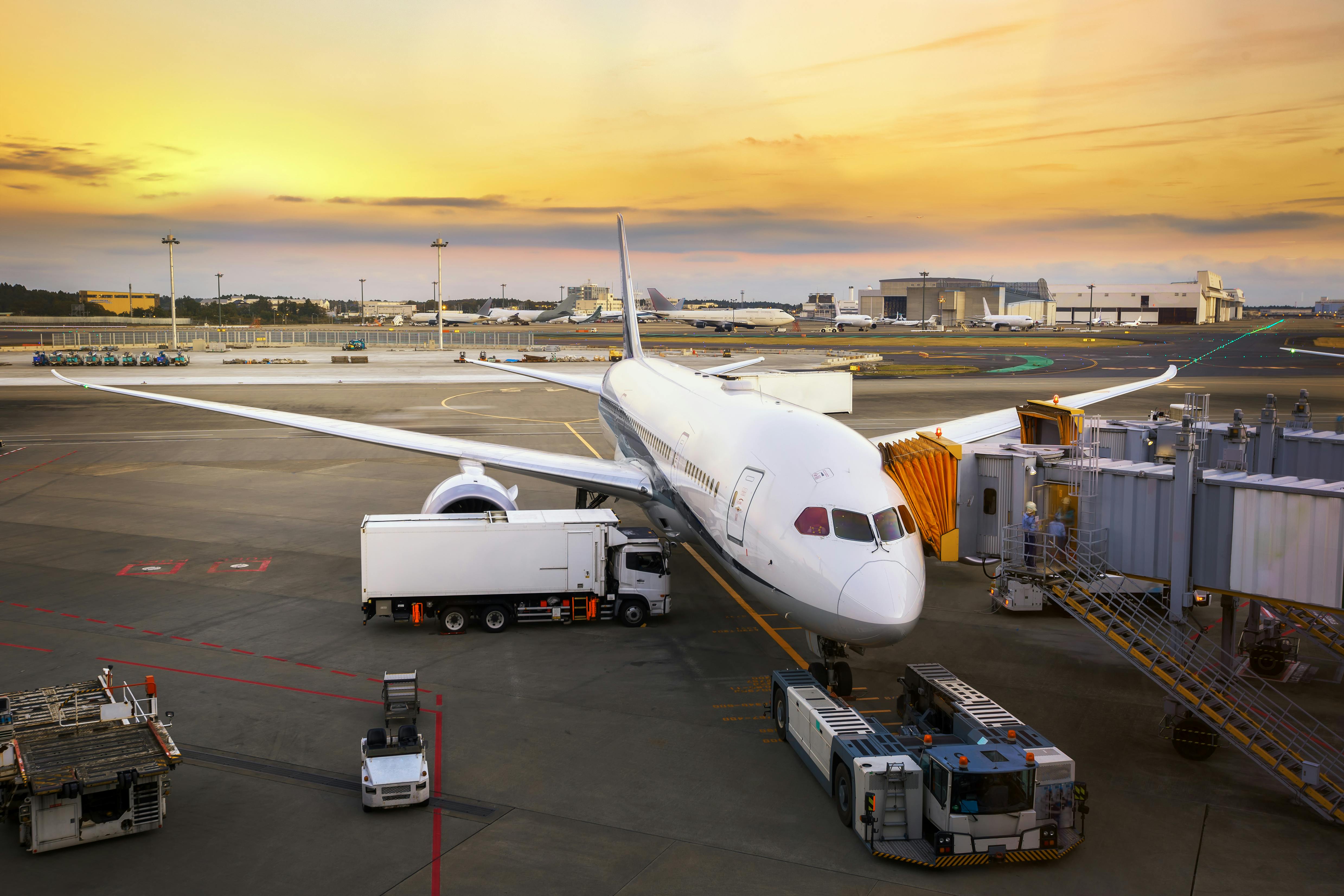 A large commercial airplane is parked at an airport gate at sunset, with service vehicles nearby, including a fuel truck and luggage carts, as it prepares for boarding on the tarmac.