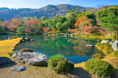 A serene Japanese garden with a clear pond reflecting colorful autumn trees, rocks, and distant wooded hills under a blue sky.