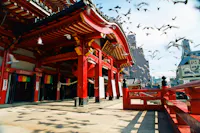 A traditional Japanese temple with vibrant red pillars, intricate wooden details, and a curved roof. Birds are flying above the temple, and sunlight casts shadows on the ground. Buildings are visible in the background.