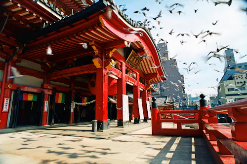 A traditional Japanese temple with vibrant red pillars, intricate wooden details, and a curved roof. Birds are flying above the temple, and sunlight casts shadows on the ground. Buildings are visible in the background.