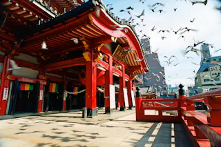A traditional Japanese temple with vibrant red pillars, intricate wooden details, and a curved roof. Birds are flying above the temple, and sunlight casts shadows on the ground. Buildings are visible in the background.