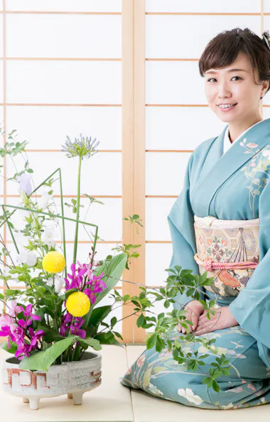 Ikebana Japanese flower arrangement A woman wearing a blue kimono sits on a tatami mat beside a traditional floral arrangement, with shoji screens in the background. She is smiling and has her hands on her lap.